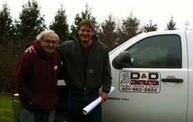 Two men standing outdoors next to a white work van with evergreen trees in the background