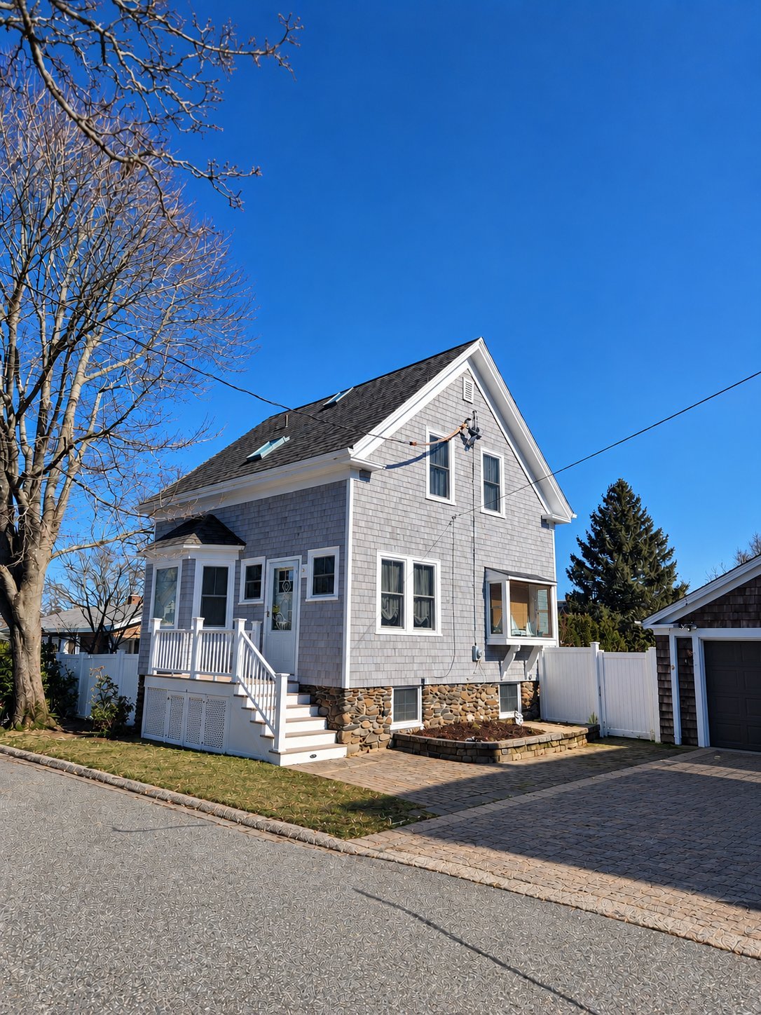Two-story white cottage-style house with front porch and stone foundation on sunny day with clear blue sky.