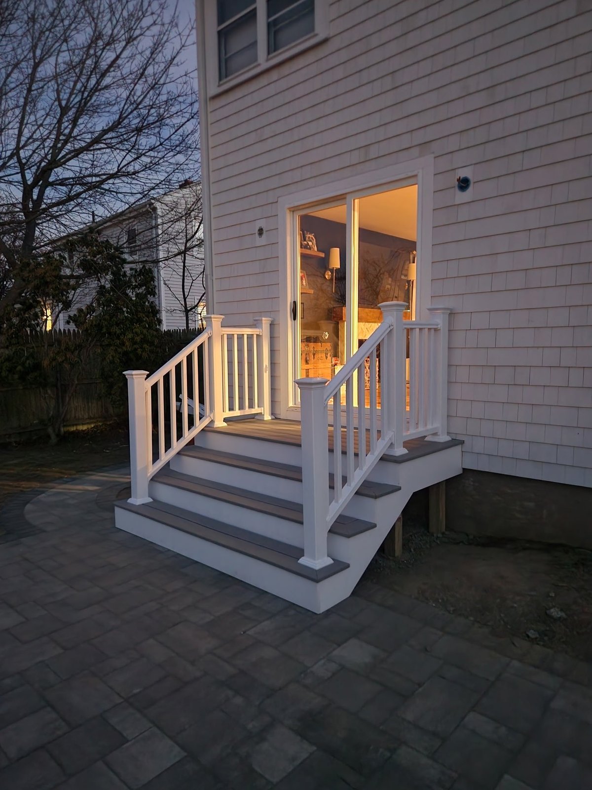White deck with stairs and railings attached to a gray house with illuminated glass doors at dusk.