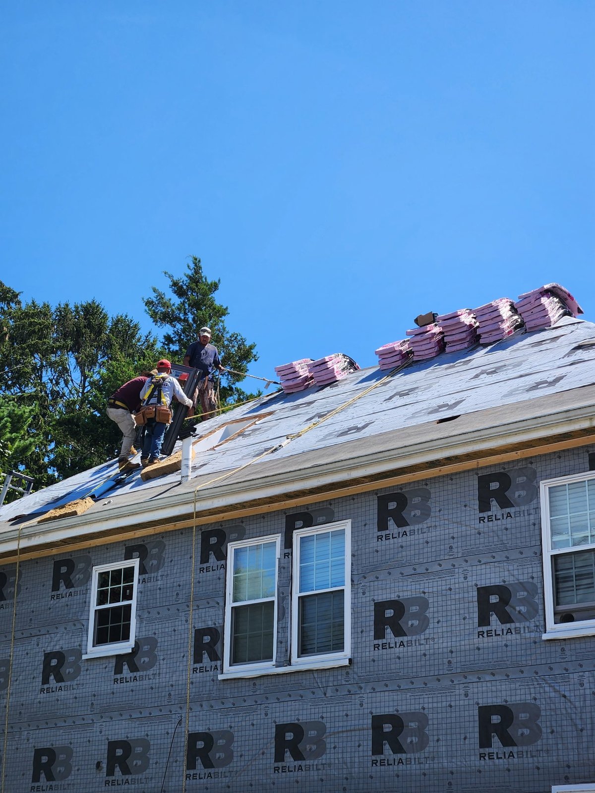 Construction worker on blue tarp installing red roof shingles on brick house under clear sky.